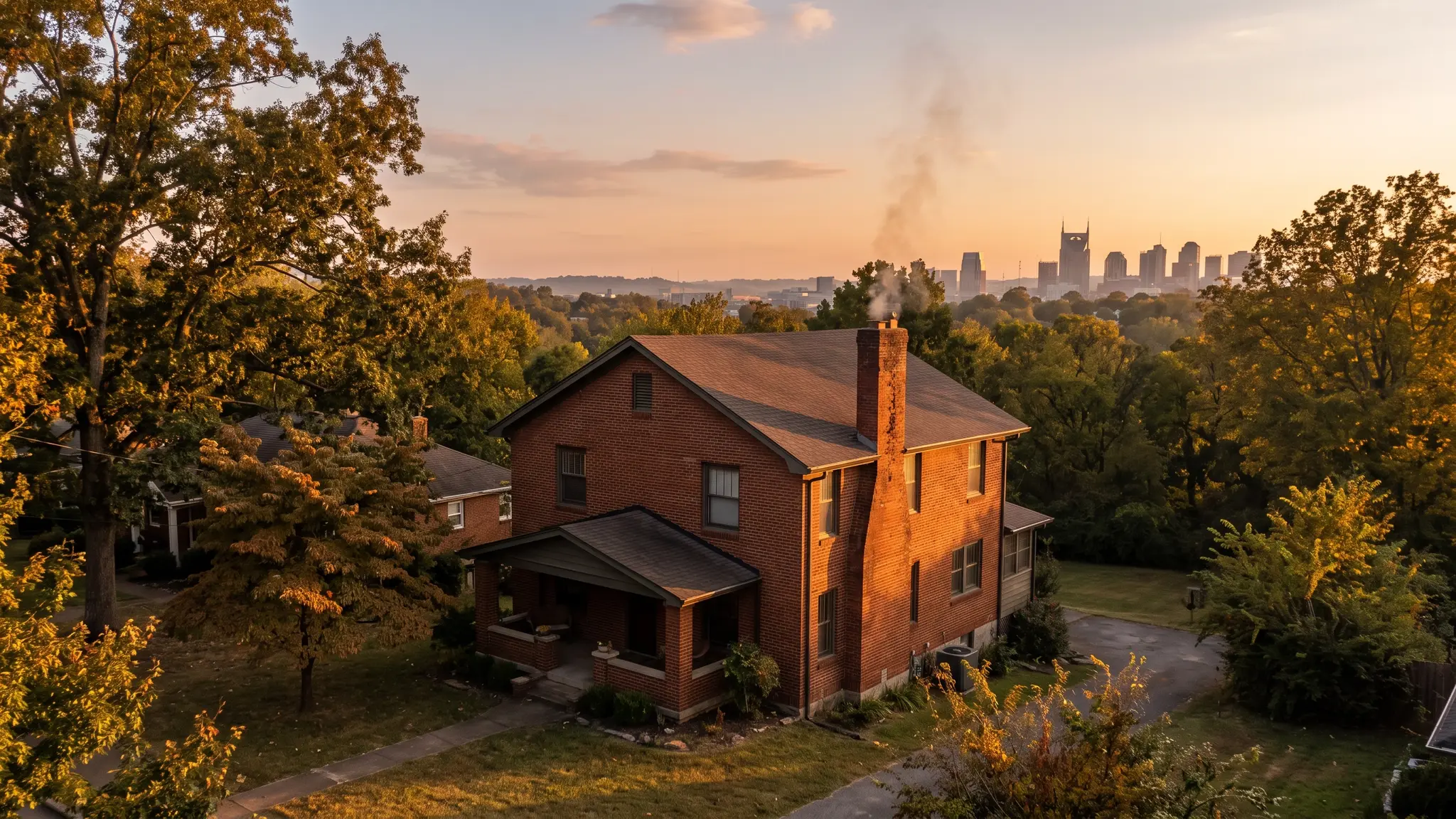 A red brick Tennessee home with a tall brick chimney releasing a thin wisp of smoke at golden hour, with the Nashville skyline softly visible in the distance